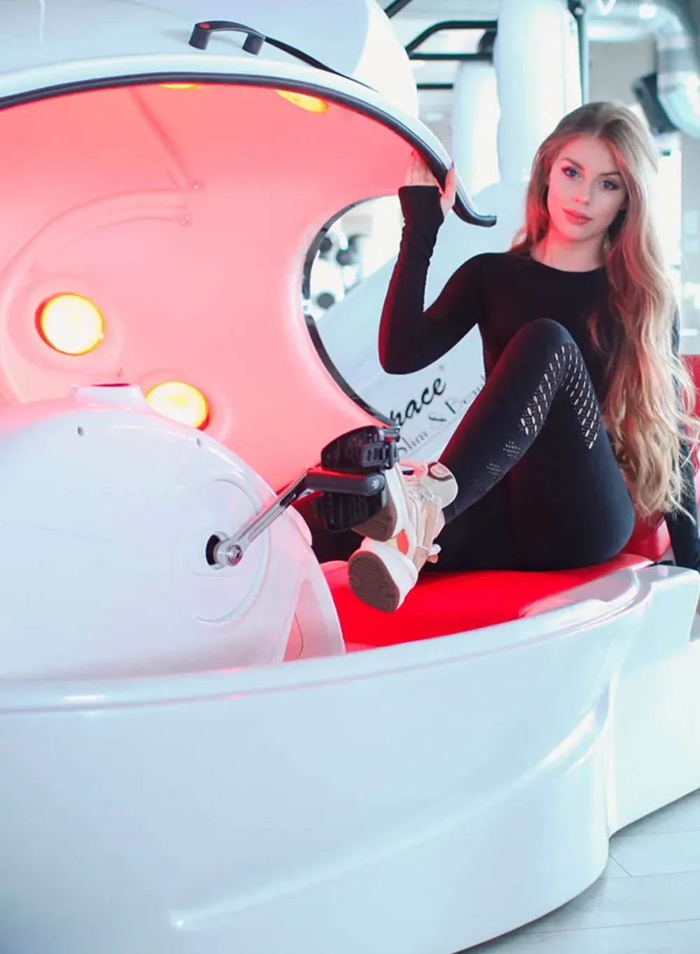 Młoda kobieta siedzi w maszynie do modelowania ciała z czerwoną iluminacją, z wyciągniętymi nogami i jedną ręką trzymającą górną część maszyny, ubrana w czarny strój i sneakersy.
