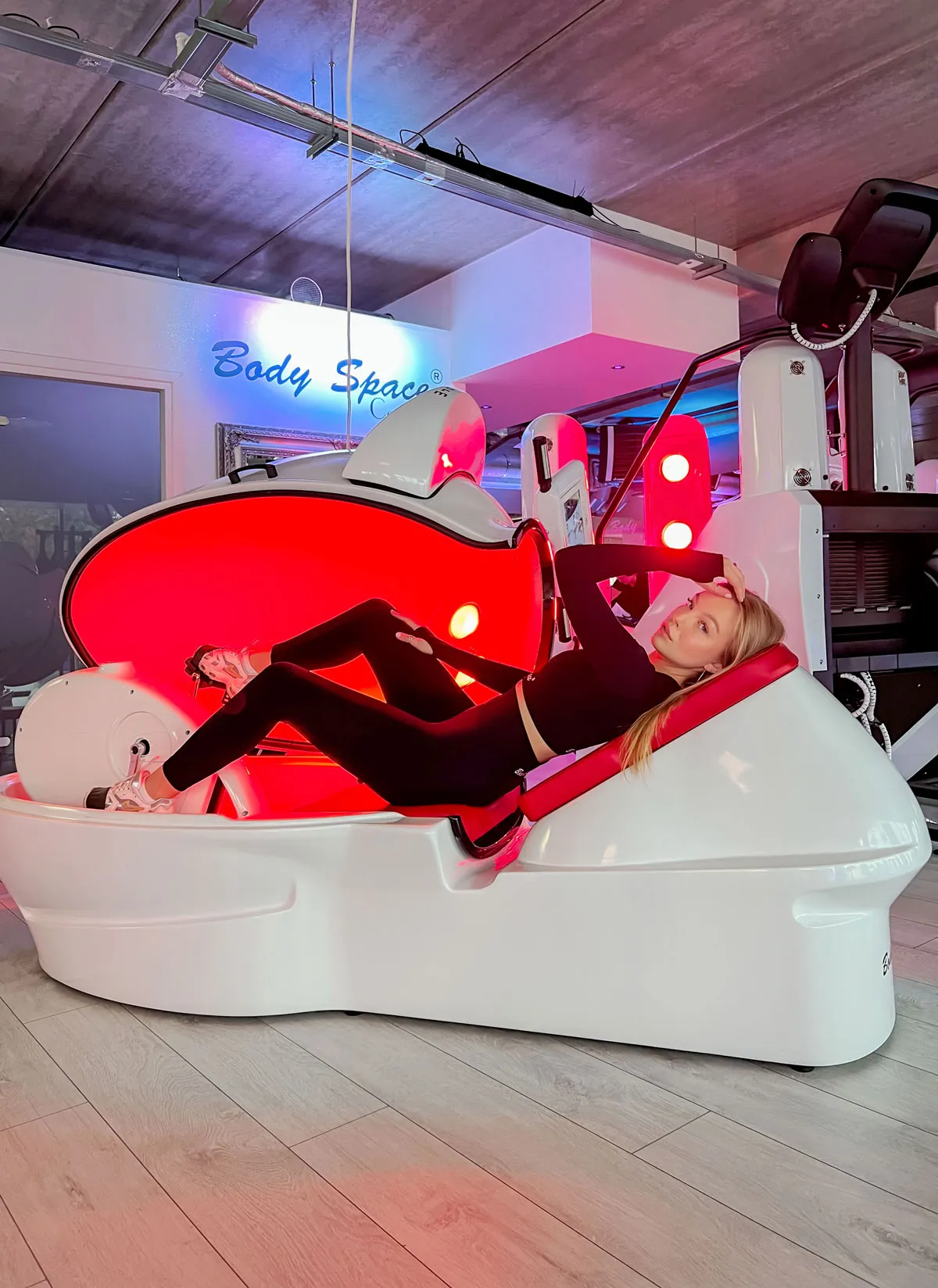 Młoda kobieta odpoczywa w urządzeniu Body Space™, leżąc na czerwono-podświetlonym fotelu w białym kombinezonie sportowym.